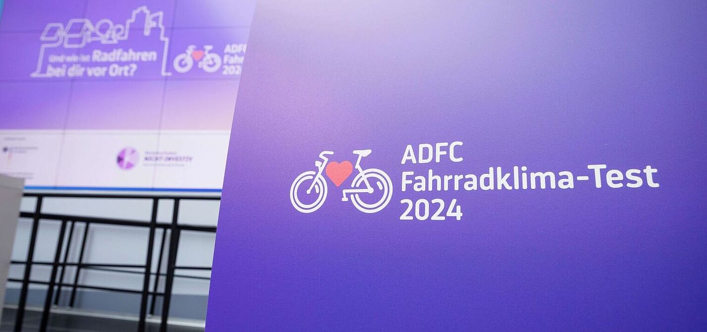 Logo des ADFC Fahrradklima-Tests 2024 auf einem violetten Pult, im Hintergrund eine Präsentationsfläche mit dem Text: „Und wie ist Radfahren bei dir vor Ort?“.