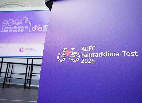 Logo des ADFC Fahrradklima-Tests 2024 auf einem violetten Pult, im Hintergrund eine Präsentationsfläche mit dem Text: „Und wie ist Radfahren bei dir vor Ort?“.